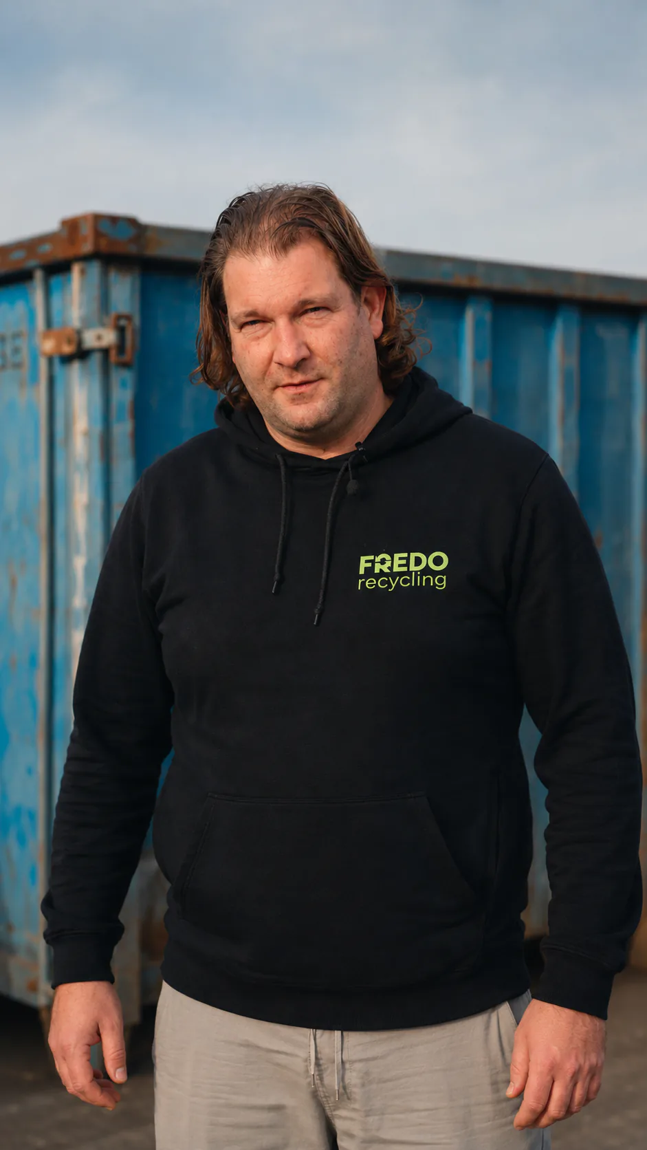 Oprichter Fredo Recycling bij schrootcontainer, Harelbeke Oprichter van Fredo Recycling in een zwarte hoodie voor een blauwe schrootcontainer op de werf in Harelbeke
